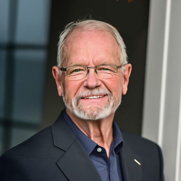 A smiling elderly man with glasses, gray hair, and a beard, wearing a dark suit and a blue shirt, standing outdoors in front of a modern building.