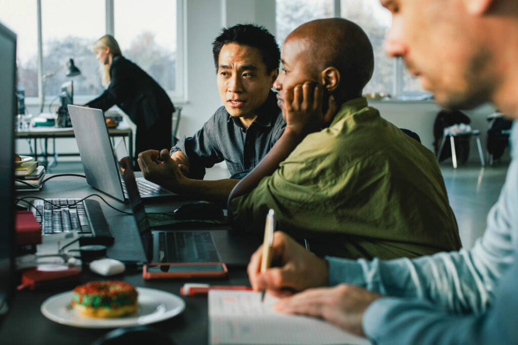 An example of a diverse group of colleagues collaborating in a modern office, with two men engaged in a discussion at a desk, one taking notes, and a woman working in the background.