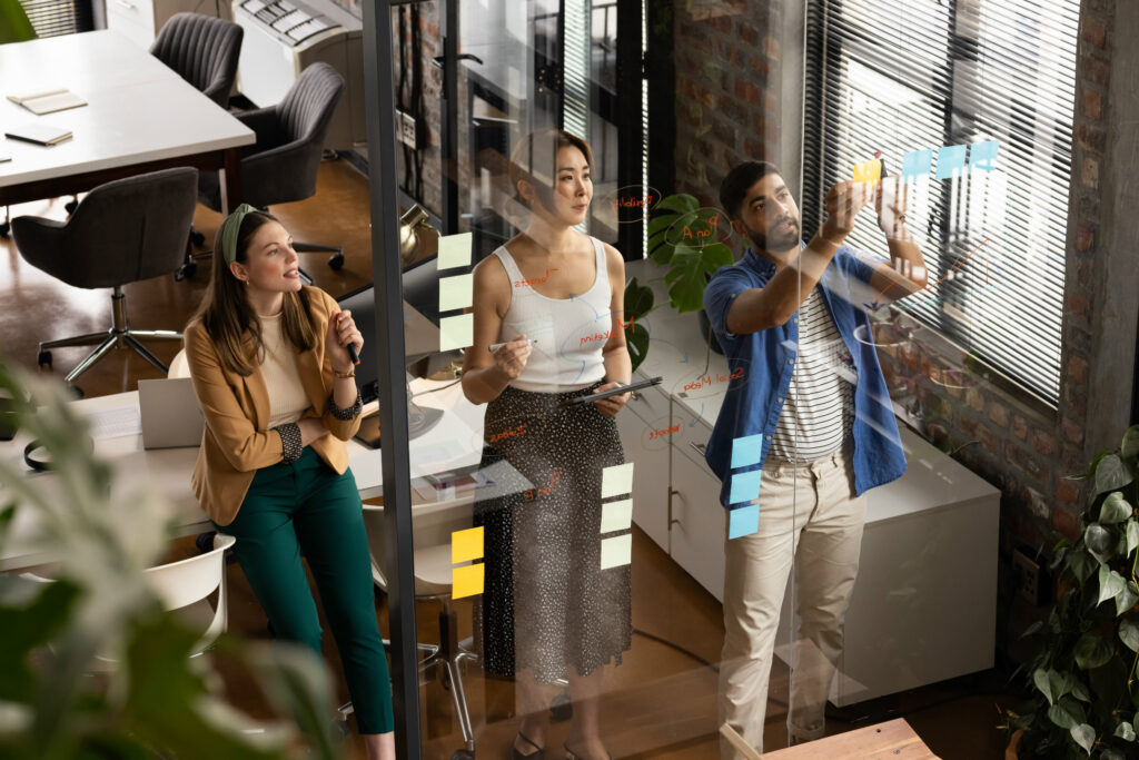 Three colleagues collaborating in a modern office, with one man writing on a glass wall with sticky notes while two women observe and discuss.