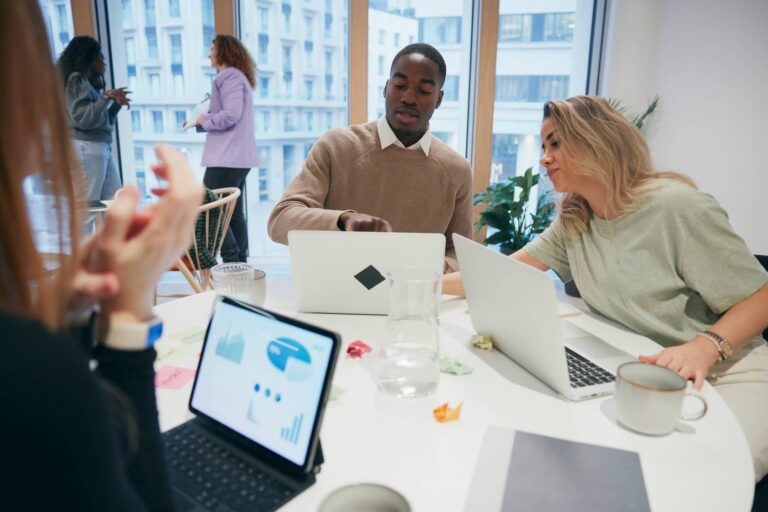 Group of young professionals working together at a bright office table with laptops and tablets.