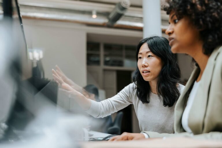 Two women engaged in a discussion in an office setting, one gesturing with her hand while the other listens attentively.
