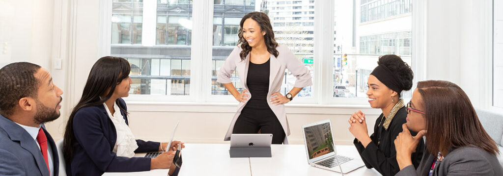 A woman standing at a conference table with four seated colleagues, in a modern office with large windows and cityscape view.