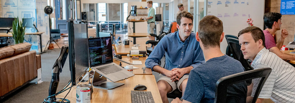 Three young men engaged in a discussion in a modern office, with computers and monitors on the desk, and a whiteboard in the background.